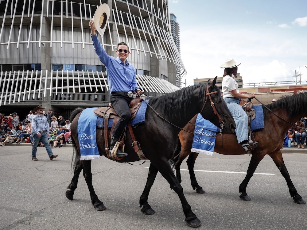 Pierre Poilievre, a 46-year-old light-skinned man, rides a dark brown horse on a city street, holding a white cowboy hat over his head.