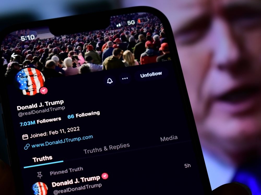 A smartphone screen displays Donald J. Trump's Truth Social page, with Trump's face out of focus in the background.