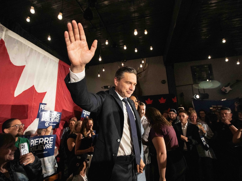 Pierre Poilievre, in a suit and tie, waves with one hand, surrounded by supporters.