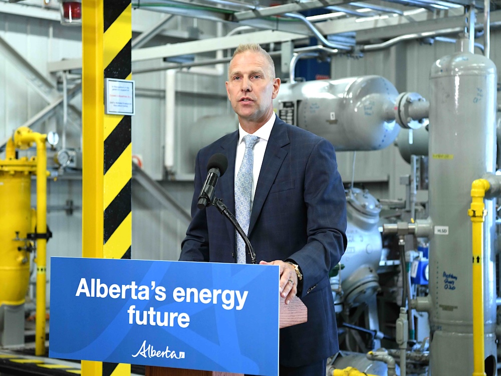 A middle-aged light-skinned man wearing a suit stands at a podium with a sign that says &lsquo;Alberta&rsquo;s Energy Future.&rsquo; Behind him are pipes and tanks.
