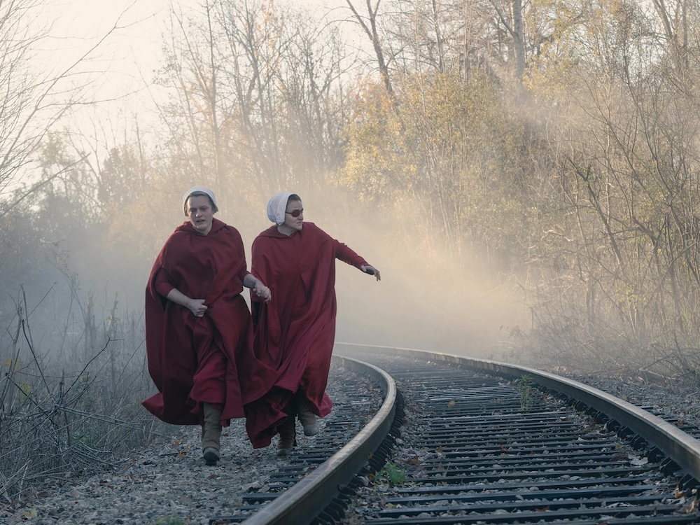 Two women in red robes and white head scarves run beside train tracks, clasping each other&rsquo;s hands.