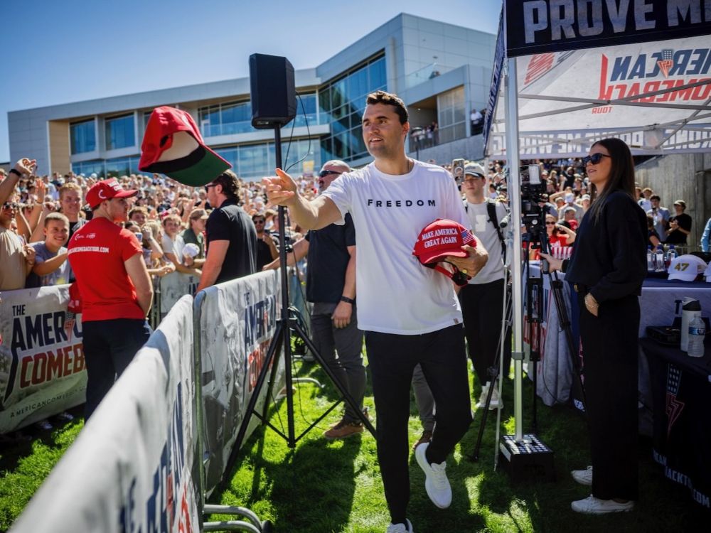 A man in his early 30s with short brown hair tosses red MAGA ball caps to university students.