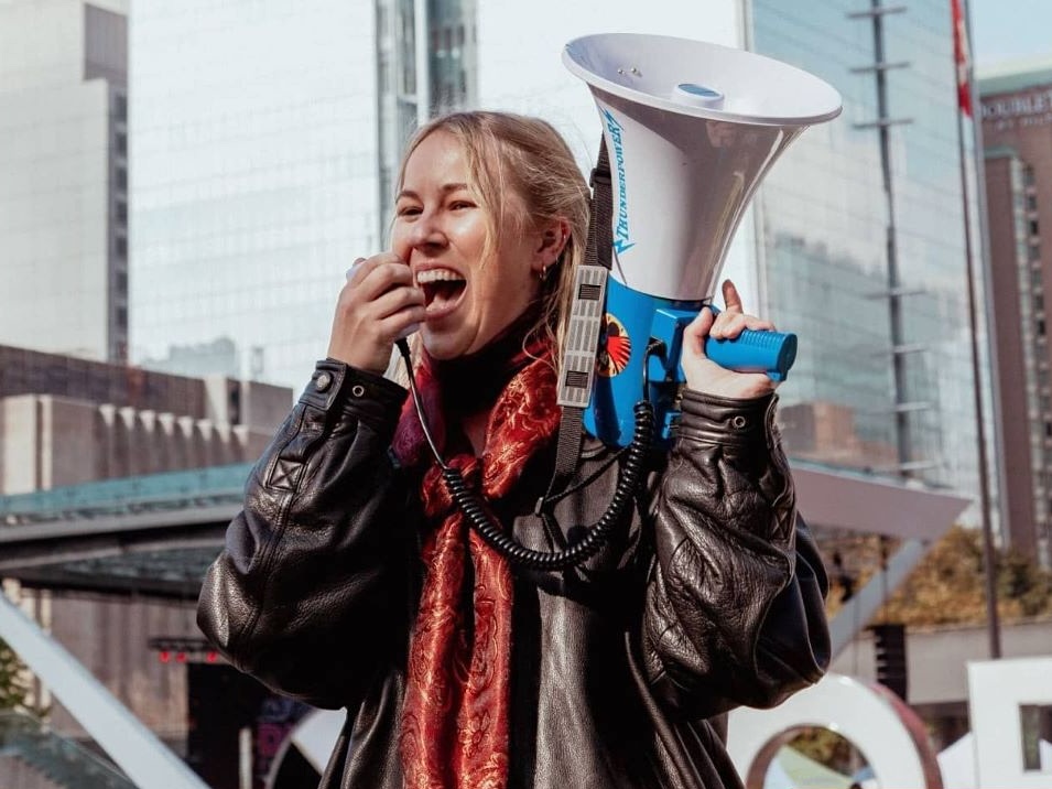 A woman with light skin and blond hair wearing a red scarf and black leather jacket speaks into a microphone attached to a white and blue megaphone.