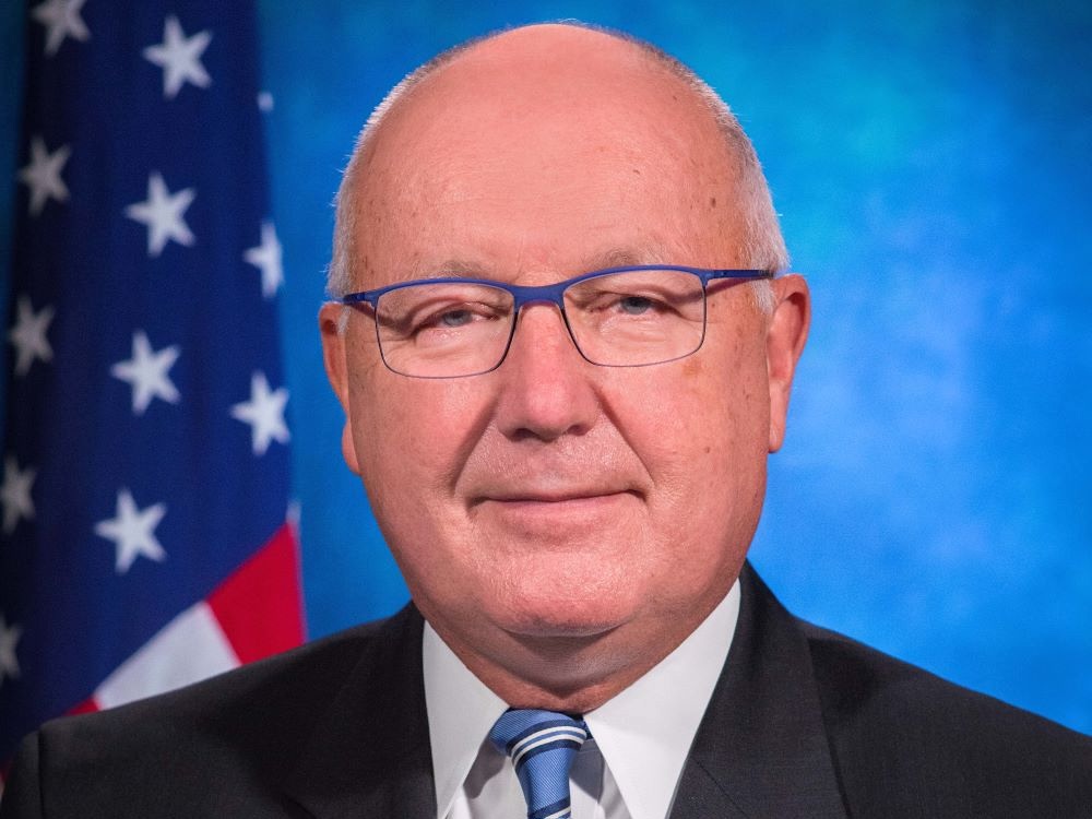 A headshot of a balding, florid white man in his 70s, wearing a suit and tie, with a blue background and the U.S. flag behind him.