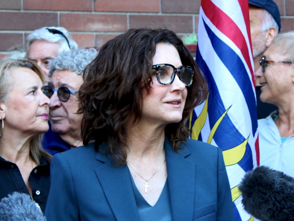 A light-skinned woman with shoulder-length brown hair stands at a podium. A group of people and the British Columbia flag can be seen behind her.