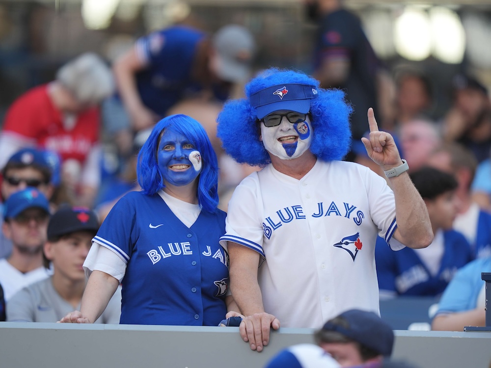 Two people are in stands, a man wearing a white Blue Jays shirt and a woman wearing a blue one. Both have their faces painted and blue hair.