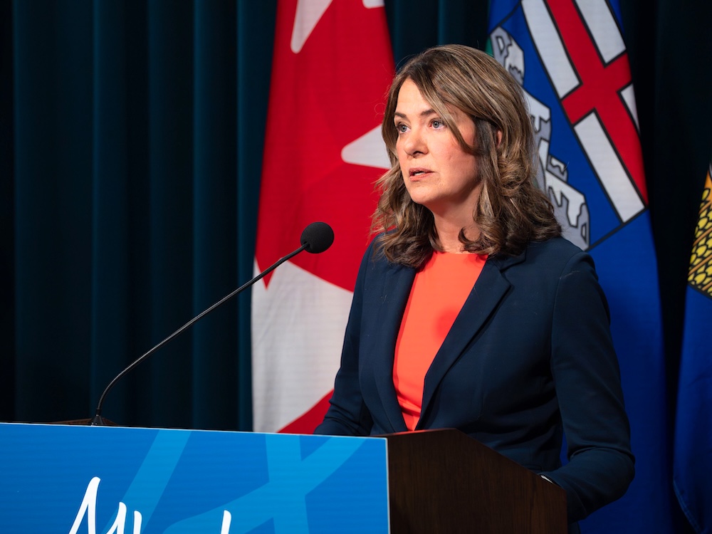 Danielle Smith, a 54-year-old light-skinned woman, stands at a lectern with Alberta and Canada flags behind her.