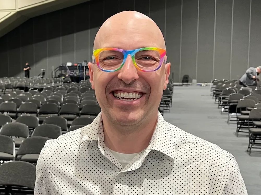 A light-skinned bald man wears a white dotted dress shirt and rainbow glasses smiles widely at the camera. Behind him are rows and rows of chairs in a large speaking hall or auditorium.