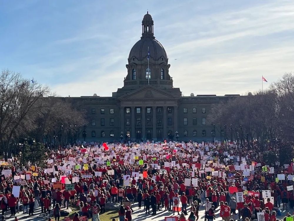 A large crowd, many wearing red and holding signs, face the Alberta legislature.