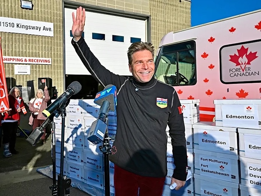 A middle-aged man with light-toned skin waves. He&rsquo;s standing in front of a wall of white cardboard boxes and an RV.