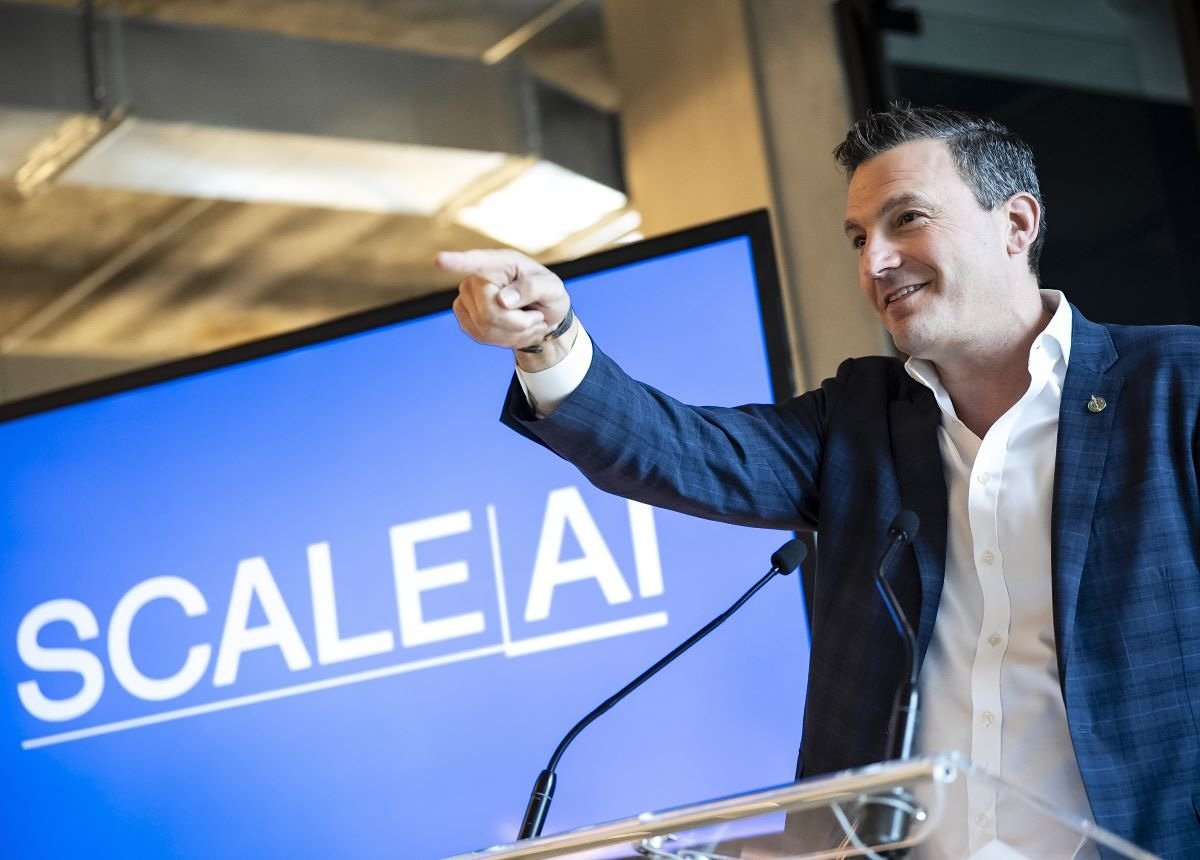 A dark-haired middle-aged man in an open-collar white shirt and blue plaid sports coat points and smiles in front of a video screen that says ‘Scale AI.’