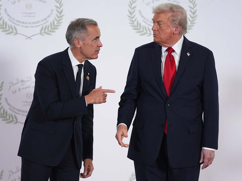Prime Minister Mark Carney leans toward Donald Trump in front of a white backdrop, pointing. Trump looks disapproving.