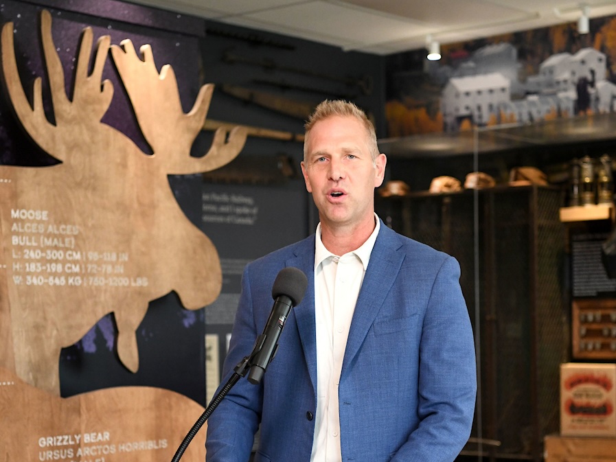 A 50-ish light-skinned man stands at a podium with a moose cutout behind him.