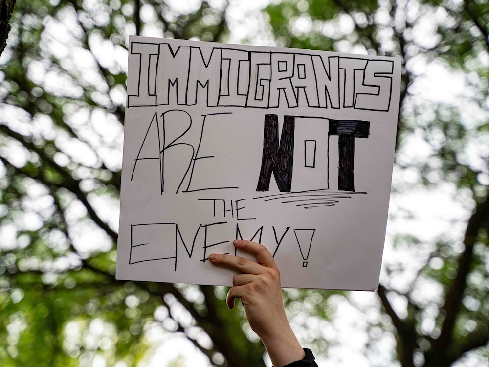 A hand holds a sign that says, &lsquo;Immigrants are not the enemy.&rsquo;