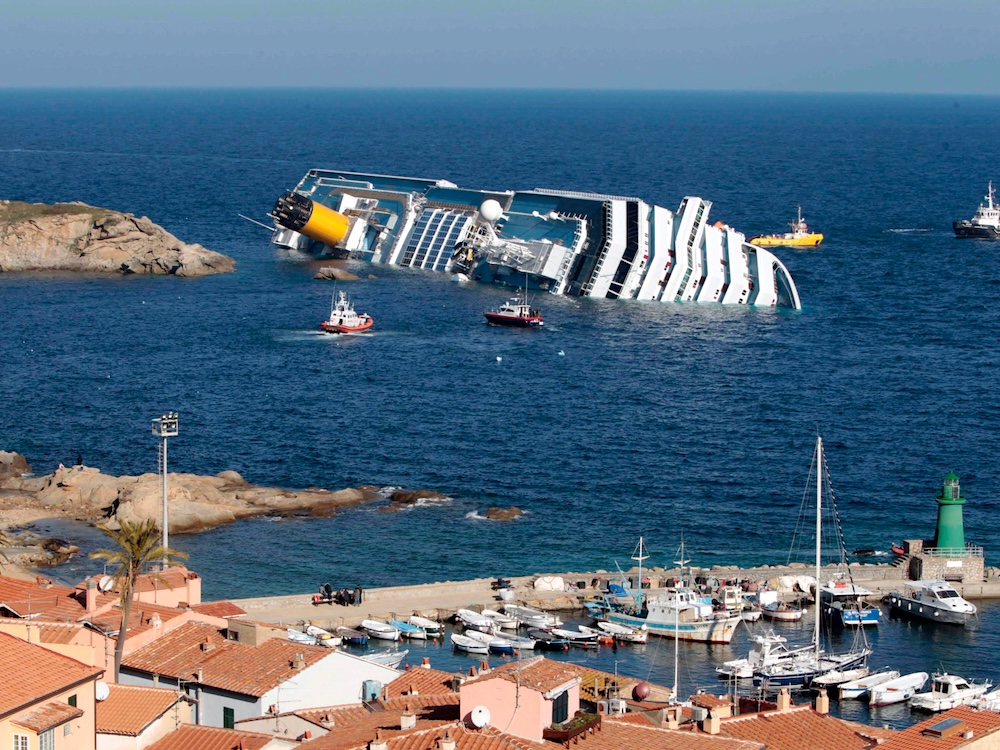 A cruise ship is on its side half-submerged in front of a village.