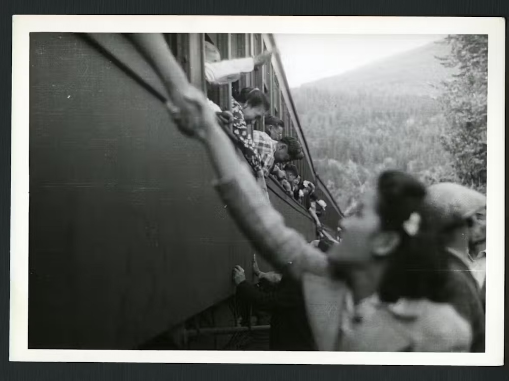 A black and white photo shows a woman reaching up to hold the hand of someone in a train.