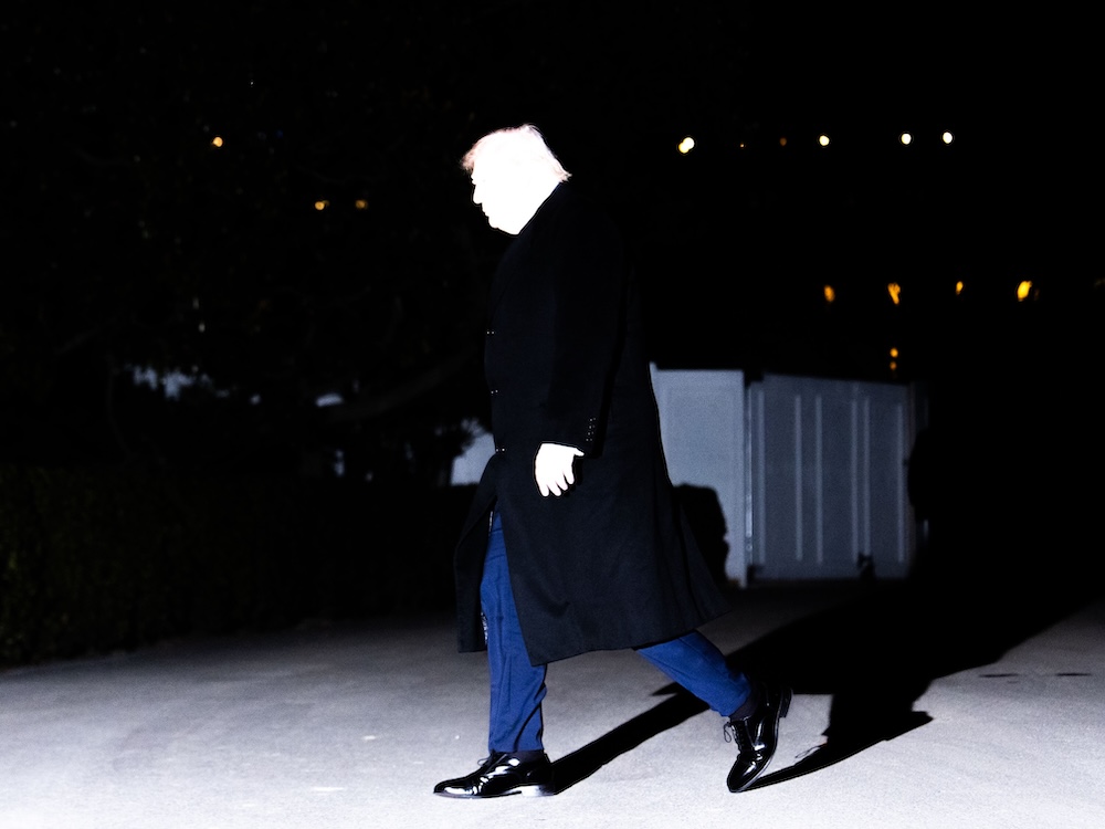 Donald Trump is shown walking. A bright flash has left his face and hand white as he wears a long dark overcoat and blue pants.