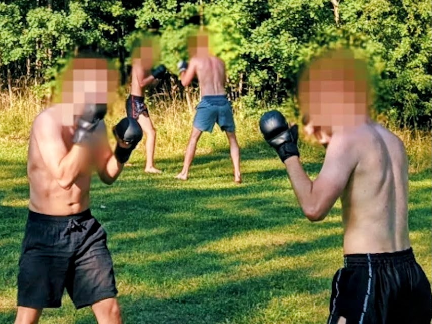 Two pairs of men, shirtless but wearing shorts and boxing gloves, train outdoors. Their faces are blurred.