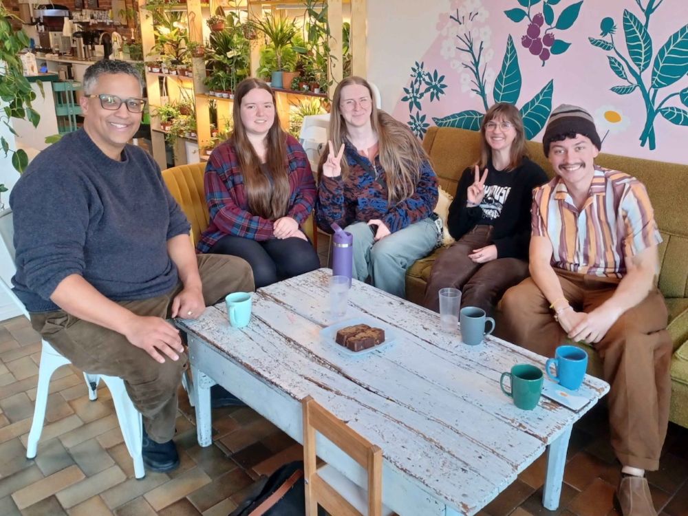 A middle-aged man meets with four people in their 20s in a coffee shop decorated with plants and a colourful floral mural.