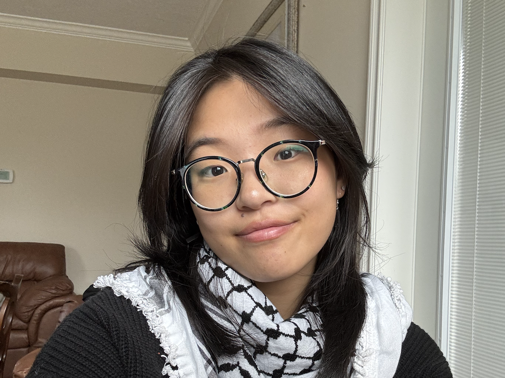 A photo of the author: a woman with medium skin tone and dark hair smiles into the camera. She's wearing glasses, a black shirt and a keffiyeh around her neck.