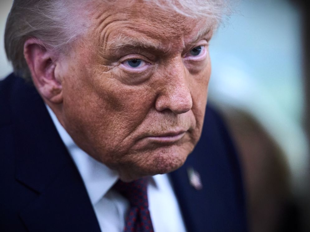 A close-up of the face of Donald Trump, who has uneven orange-tinted skin and silver-white hair and wears a navy blue suit.