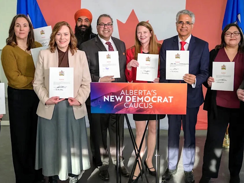 Naheed Nenshi and seven other NDP MLAs stand in front of a Canadian flag, holding pieces of paper.