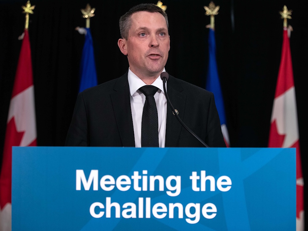A light-skinned man in his mid-40s, wearing a suit and with short dark hair, stands at a podium with a sign that says, &lsquo;Meeting the challenge.&rsquo;