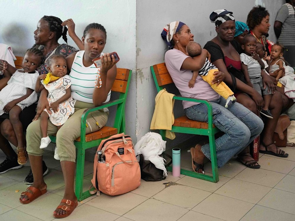 Women sit on wooden benches with green trim in a crowded indoor space with tile floors. Many are holding small children in their arms or laps.