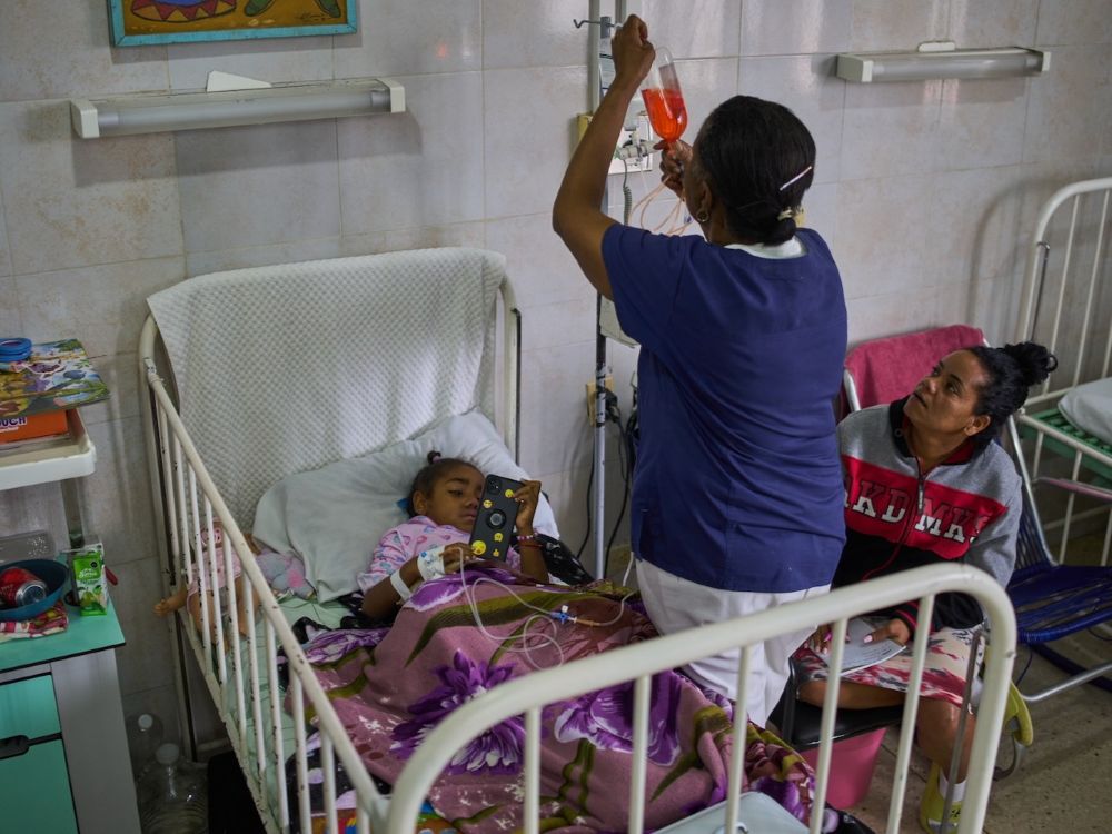 A little girl lies in a hospital bed with high guard rails as her mother sits in a chair by her bed. A man is adjusting the patient&rsquo;s IV.