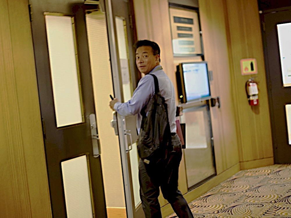 Mayor Ken Sim, wearing a button-down shirt and carrying a shoulder bag, looks over his shoulder as he opens a door at city hall.