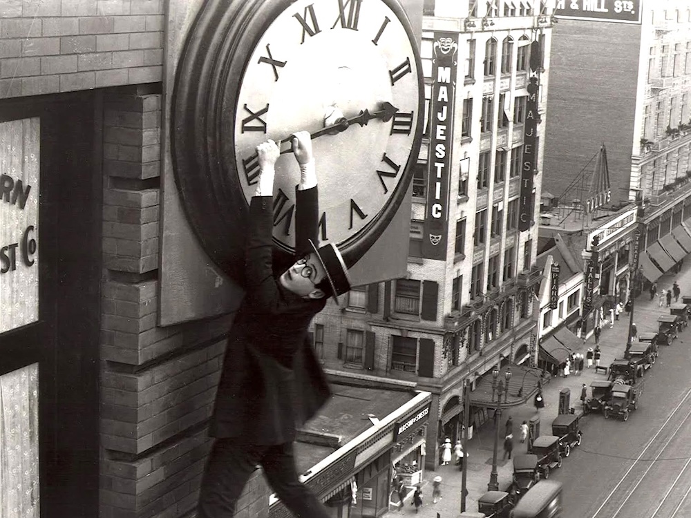 A black and white image from the 1920s shows a bespectacled man in a dark suit and boater hat hanging from the hands of a clock high above a city street.