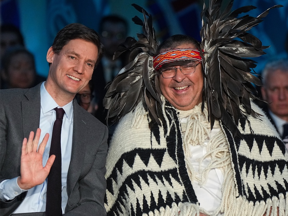 A middle-aged man with short brown hair and wearing a grey suit waves and smiles. He sits beside another man who is dressed in Musqueam regalia, including a headpiece with large feathers and a woven black and white cloak.