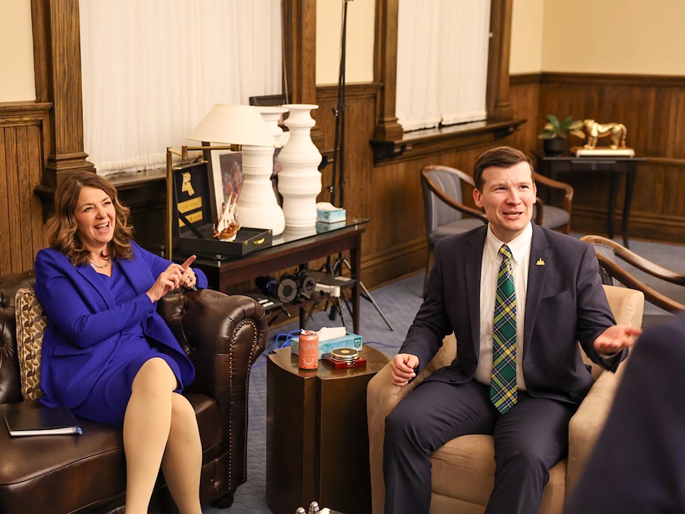 Danielle Smith and Jeromy Farkas sit an office. A golden cat is on a table in the background.