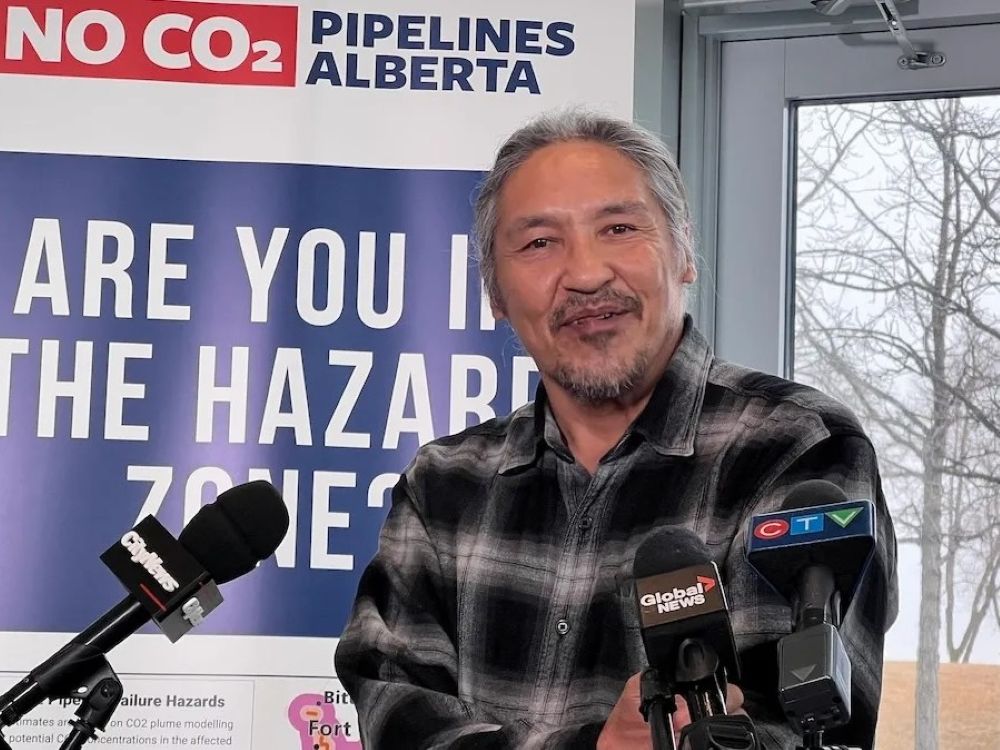 A smiling Indigenous man with grey hair and a short beard has his arms crossed. Behind him a sign says &ldquo;No CO2 pipelines Alberta&rdquo; and &ldquo;Are you in the hazard zone.&rdquo;