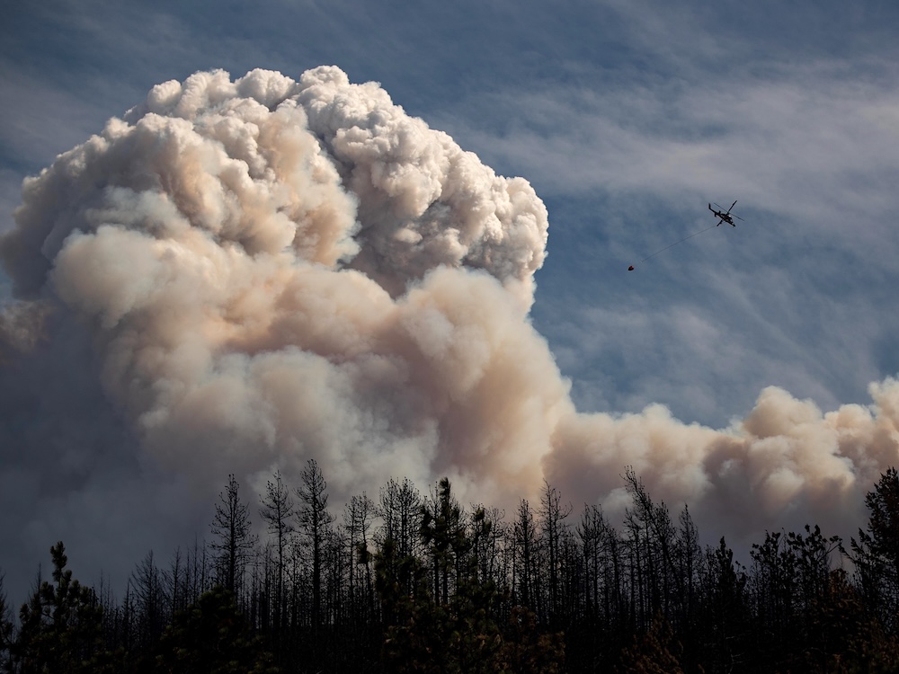 A helicopter carrying a canvas water bucket flies beside a giant cloud and smoke.