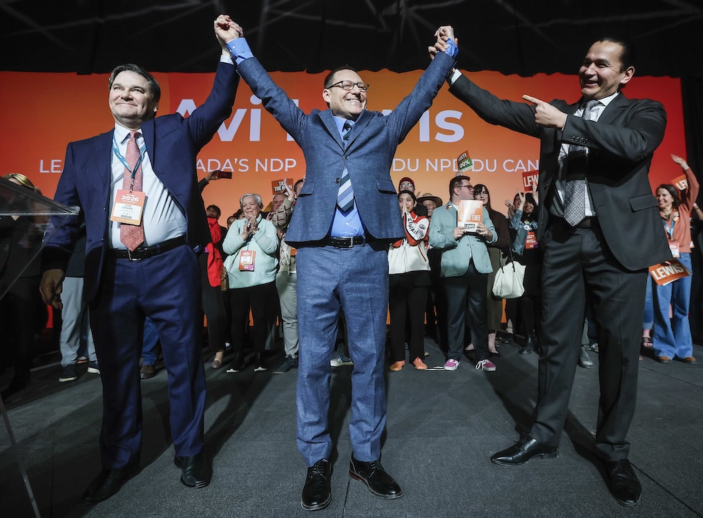 Three middle-aged men stand on a stage with supporters in background. They all wear suits and are clasping upraised hands.