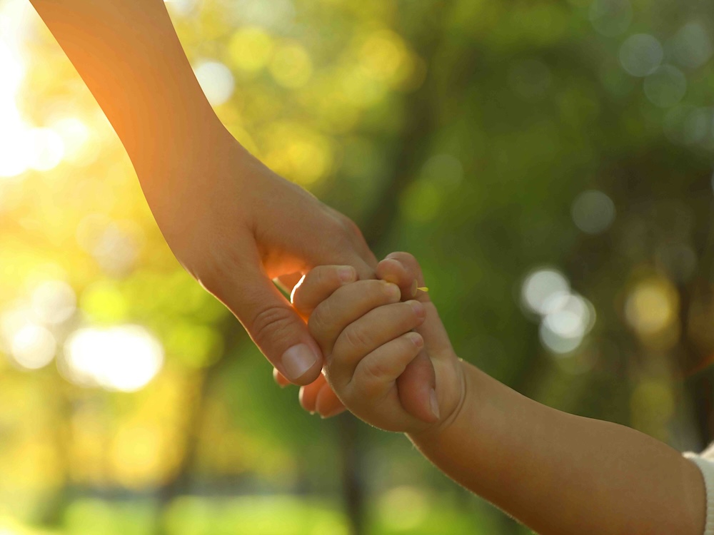 An unseen adult holds the hand of an unseen child against a bright green background.