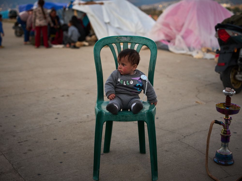 A little boy wearing a grey sweatsuit that says &lsquo;Hello&rsquo; sits in a green plastic chair in a parking lot, with plastic-covered tents in the background.