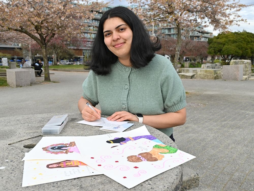 A teenage girl with medium-light skin tone and dark shoulder-length hair, Emaan Khalid, smiles as she writes on a card. In the foreground are some other cards that she has crafted laid out on a stone park table. In the background is the rest of the park, with trees colourful and in bloom. 