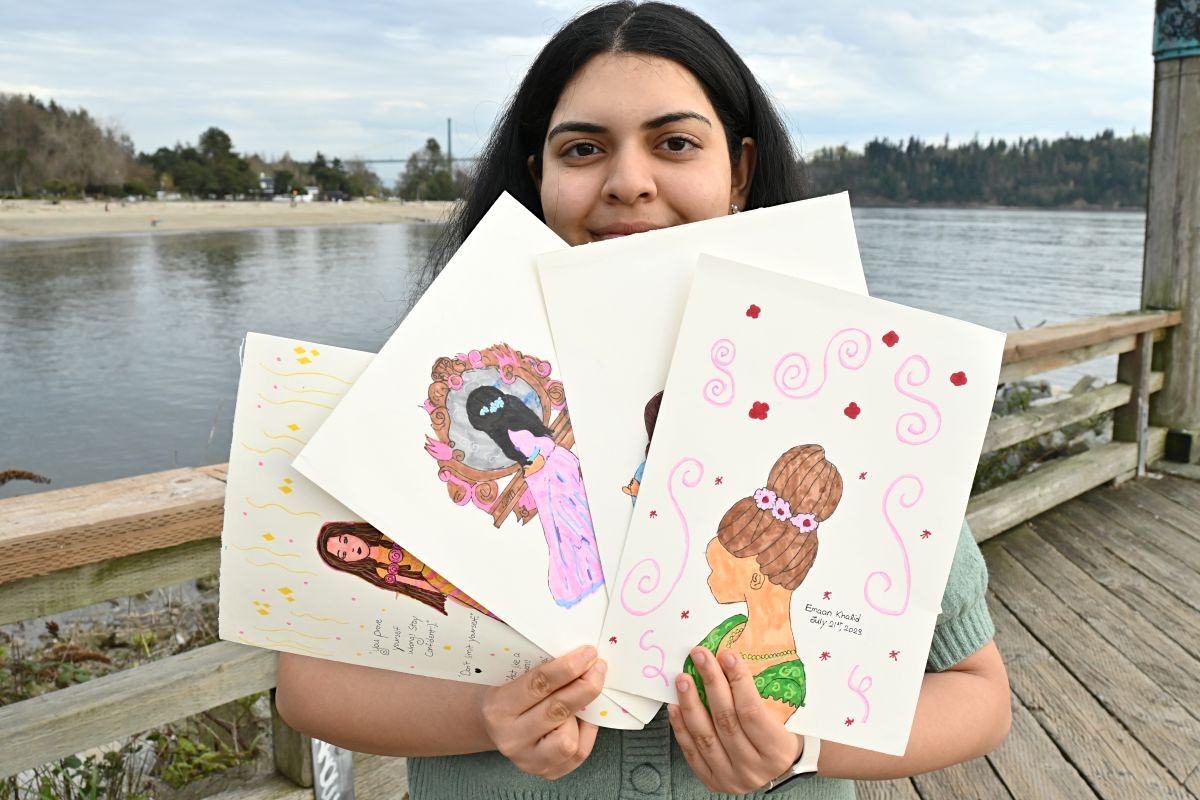 A teenage girl with medium skin tone and dark shoulder-length hair, Emaan Khalid, holds several cards spread out in front of her. On the cards are various colourful drawings of figures in elegant hairstyles and dresses.