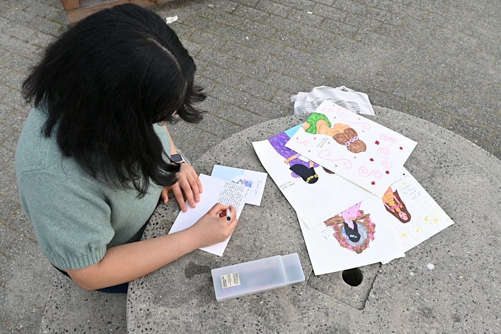 An overhead view of a dark-haired teenager, Emaan Khalid, writing a card on a stone table, with other hand-drawn cards on the table beside her.