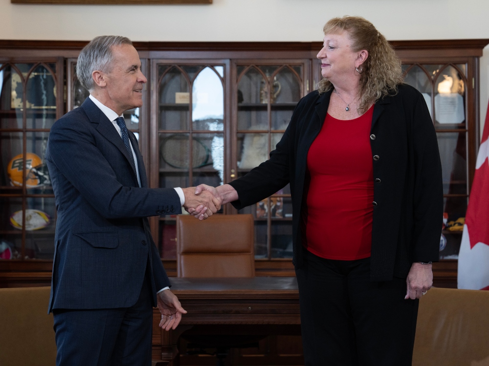 A man wearing a suit shakes the hand of a taller woman wearing a blazer and a red shirt in a government building.