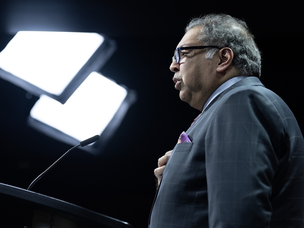 A 54-year-old man with medium skin tone stands in profile in front of a black background with bright square television lights.