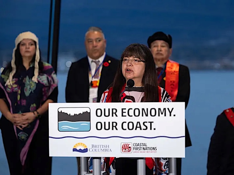 A middle-aged Indigenous woman stands at a podium that says &lsquo;Our Economy, Our Coast&rsquo; with other Indigenous people behind her.