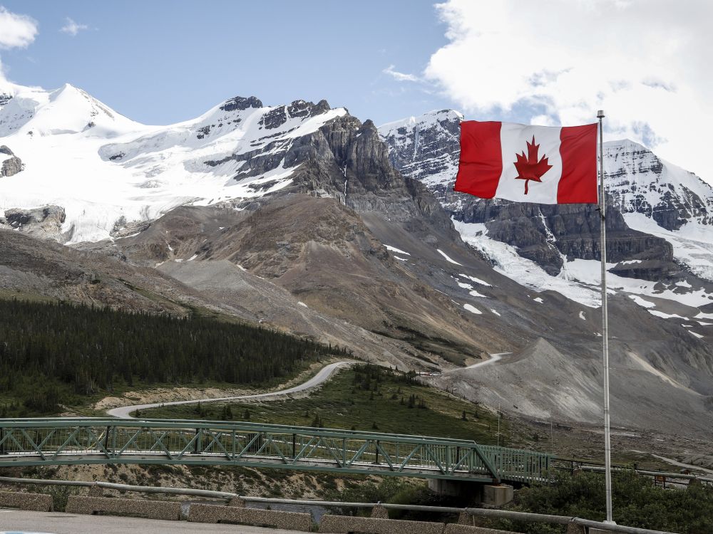 A red and white Canadian flag flies in the foreground. In the midground is a bridge leading to a hilly forest road. The background is the snow peaked mountains of the Rockies. 