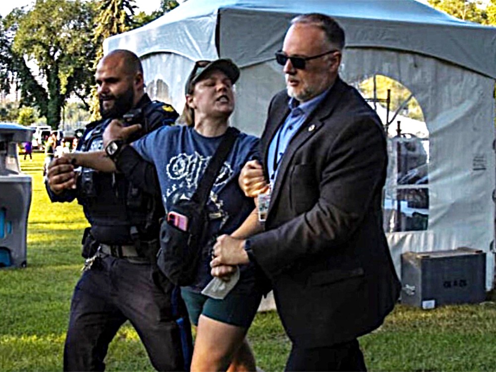 A young light-skinned woman wearing a ball cap and blue T-shirt is taken away by two large men holding her arms.