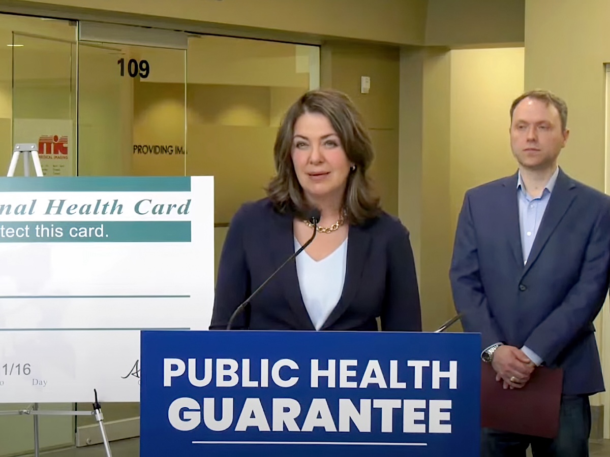 Alberta Premier Danielle Smith, a middle-aged white woman with shoulder-length brown hair, wearing a suit, stands behind a dais with a sign on it saying &lsquo;Public Health Guarantee.&rsquo;