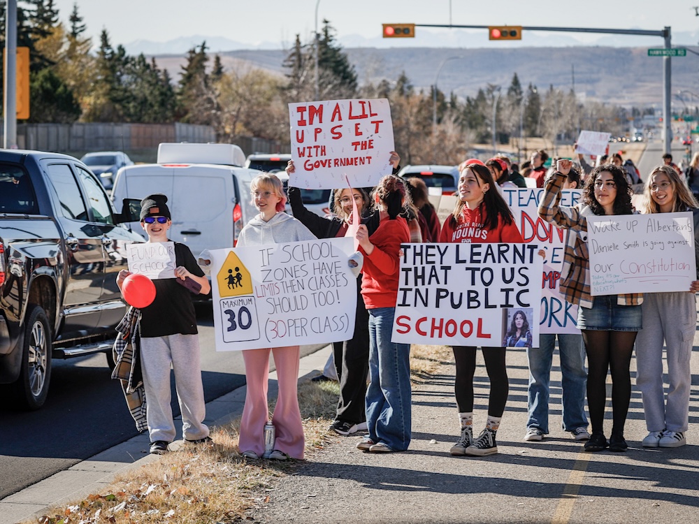 A group of striking teachers stand on a sidewalk holding protest signs.