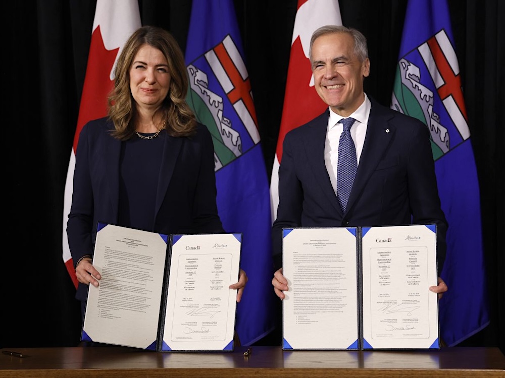 Premier Danielle Smith, a white woman with shoulder-length dark hair, and Prime Minister Mark Carney, a grey-haired white man, each smile and hold documents.