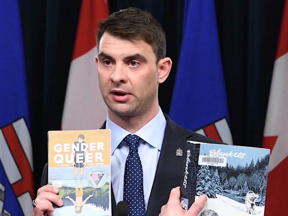 A 40-ish man with light-toned skin and dark hair is wearing a suit and holding two books titled &lsquo;Gender Queer&rsquo; and &lsquo;Blankets.&rsquo;